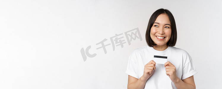 钱表情摄影照片_微笑的韩国女性展示信用卡，脸上挂着幸福的表情，站在白色背景的 T 恤上