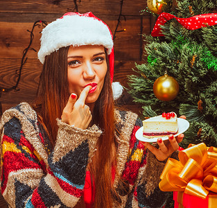 聪明的女士拿着一个盘子，里面放着一块蛋糕，嘴里放着手指，平安夜，圣诞晚餐，圣诞蛋糕，圣诞树，圣诞礼物，木头背景。