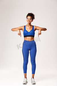 蓝色运动服摄影照片_穿着蓝色运动服的快乐非洲裔美国健身教练，指指点点，微笑，推荐健身装备，展示标志或运动宣传片，白色背景