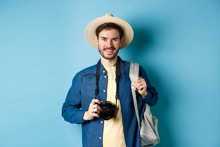 快乐的帅哥去度假，戴着夏日帽子，背着带相机的背包拍照，微笑着度假兴奋，站在蓝色背景上