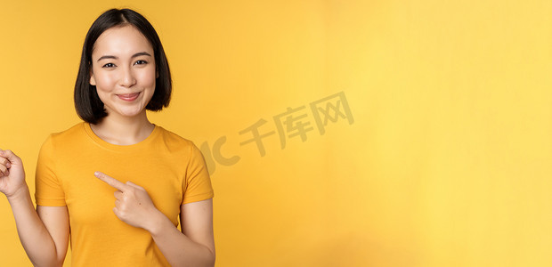 微笑的亚洲女性手指向左，在空的复制空间上展示广告，站在黄色背景上