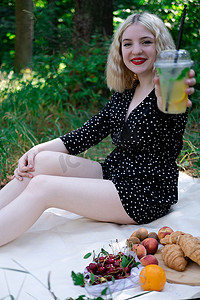 漂亮的花纹摄影照片_漂亮的金发女孩在公园的白色格子上野餐，手里拿着一个装有柠檬水的透明塑料杯。