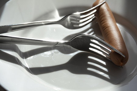 一根香肠被刺穿在白色盘子上的两个叉子上，阳光洒落。