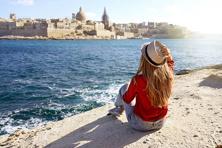 戴着帽子的美丽年轻女子坐在海边的石头上，欣赏马耳他瓦莱塔市令人惊叹的全景