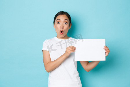 穿着白色 T 恤的亚洲女孩惊奇而兴奋，发布公告，在展示标志时惊奇地喘着粗气，一张空白纸上贴着你的广告，站在浅蓝色背景中