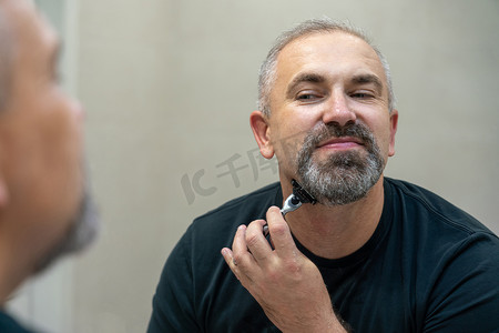 剃胡子的男人摄影照片_迷人的中年男子在镜子前剃掉胡子和胡子，让自己看起来更年轻