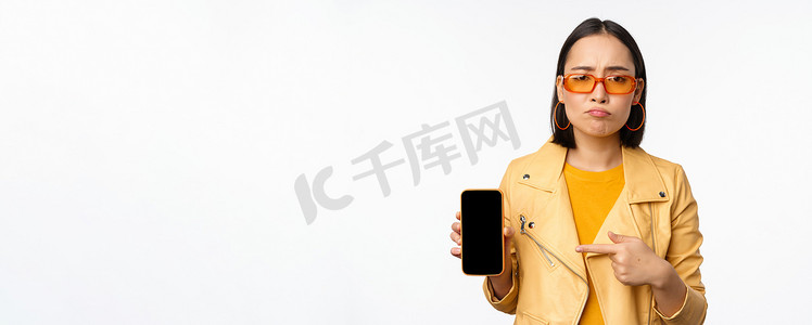 戴墨镜的悲伤亚洲女性肖像，手指指向手机应用程序界面，展示智能手机应用程序，站在白色背景上