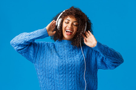腰部肖像无忧无虑的快乐微笑的女人放松，一起唱歌，在耳机中玩卡拉 OK 应用程序，闭上眼睛热情地对着耳机中的音乐进行口型同步，站在蓝色背景