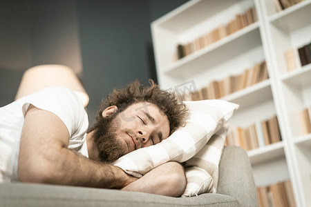 工作生活睡觉摄影照片_长时间工作到深夜后，睡在沙发上的留着胡子的英俊年轻人抱着枕头。