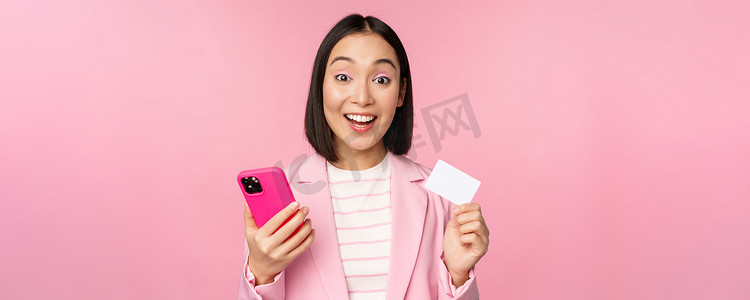 微笑快乐的亚洲女商人展示信用卡、在智能手机应用程序上在线支付、用手机订购、站在粉红色背景下的图片
