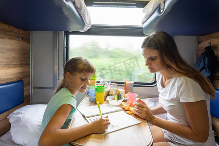 火车发展摄影照片_妈妈和女儿坐在火车的桌子旁，解决儿童杂志上的难题