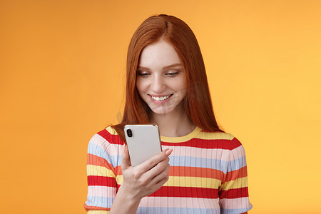 拿着照片摄影照片_迷人的现代红发女大学生拿着智能手机查看消息框看起来很开心，微笑着高兴的手机显示屏在网上收到数百张喜欢的照片，橙色背景