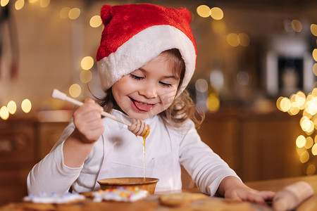 吃蜂蜜的红色圣诞老人帽子的小女孩使用蜂蜜棒 仙女灯装饰的厨房。