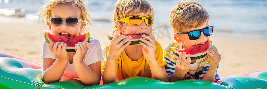 西瓜大海摄影照片_孩子们戴着太阳镜在沙滩上吃西瓜横幅，长版