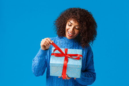 好奇而快乐、微笑的非洲裔美国女人，穿着冬衣的生日女孩，拉着礼物结打开礼物，看看里面有什么，庆祝圣诞节，新年假期，蓝色背景