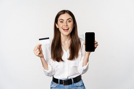 购物app摄影照片_快乐的女人展示信用卡和智能手机屏幕，网上购物的概念，在应用程序中购买，站在白色背景上