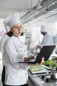 比赛工作人员摄影照片_餐厅厨房工作人员用笔记本电脑搜索美食晚餐菜肴，用于餐厅举行的烹饪比赛。