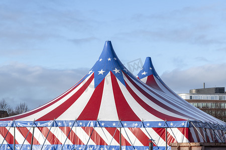马戏团帐篷摄影照片_红色和白色的马戏团帐篷，顶部覆盖着蓝色的星星盖，在阳光明媚的蓝天和云彩的衬托下