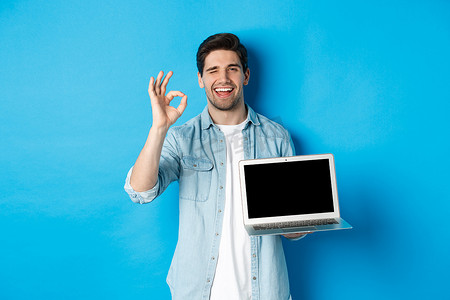 年轻人展示笔记本电脑屏幕和好的标志，批准或喜欢互联网上的促销，满意地微笑，站在蓝色背景上