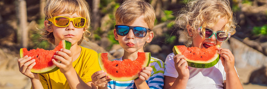 孩子们戴着太阳镜在海滩上吃西瓜横幅，长格式