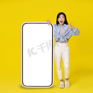 兴奋的亚洲女孩竖起大拇指，靠在巨大的、巨大的智能手机上，上面有白色、空白的屏幕，靠在黄色背景中孤立的休闲装上。