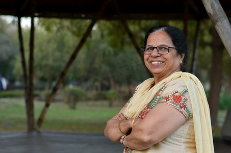 印度新德里摄影照片_夏季，印度新德里，聪明而自信的北印度高级女性站在公园里，双手交叉/折叠，在镜头前摆出姿势，穿着灰白色萨尔瓦·卡米兹旁遮普套装