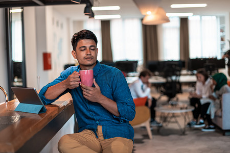 在现代开放式创业办公室的休闲区喝茶时，休闲商人使用笔记本电脑休息