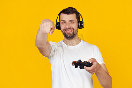 穿着白色 T 恤留着胡子的年轻人微笑着，游戏玩家戴着耳机玩电子游戏，用手指指着相机和你，手势，正面和自信的手势。