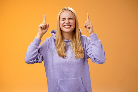 满怀希望的迷人金发女学生祈祷赢得一等奖实现梦想终生希望财富她身边闭上眼睛微笑激动向上期待好消息站立橙色背景