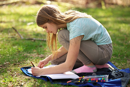 漫画美女涂色摄影照片_一个年轻的少女在草地上的毯子上的笔记本或日记上写字或涂色
