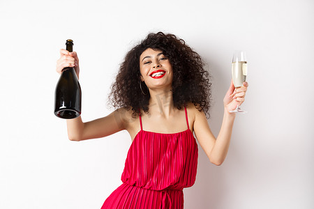 穿着红色裙子的快乐派对女孩，拿着一瓶香槟和玻璃杯跳舞，喝酒玩乐，庆祝节日，站在白色背景上