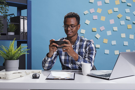 工作玩手机摄影照片_非洲裔美国商人在现代工作空间办公室里用移动智能手机玩游戏。