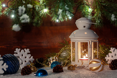 雪中​​矗立着白色灯笼，上面有燃烧的蜡烛，周围是木墙、圣诞树枝和灯光的圣诞装饰品