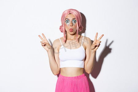 戴着粉红色假发的傻女孩的形象，庆祝万圣节，展示滑稽的面孔和粘着的舌头，做和平标志，站在白色背景上