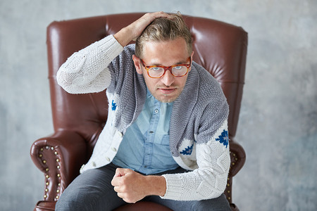 一个戴眼镜的时尚聪明男人的肖像凝视着镜头，视野开阔，身材矮小，胡子拉碴，魅力四射，蓝色衬衫，灰色毛衣，坐在棕色皮椅上