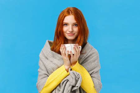 可爱、浪漫、漂亮的红发女性，戴着灰色围巾，变暖，喝热茶，拿着咖啡，微笑的镜头高兴而放松，站在蓝色背景上高兴