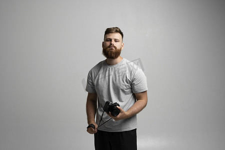 带单反相机的留着胡须的专业摄影师的肖像直视灰色背景中隔离的相机。