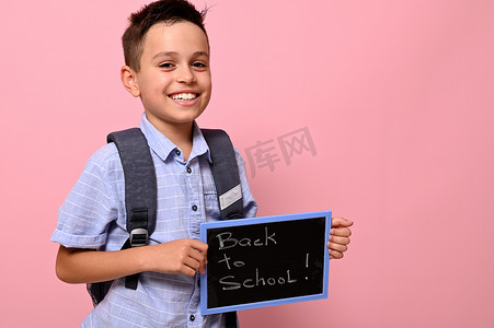 粉笔字学习摄影照片_背着背包的可爱小学生拿着黑板，上面写着粉笔字，回到学校，对着镜头露出可爱的微笑，在粉色背景中被隔离，有文字空间