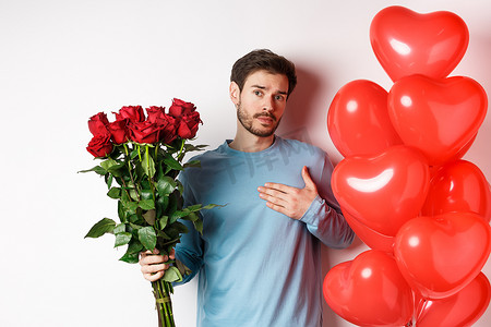 浪漫的男人在情人节用礼物表达他的爱，带来一束红玫瑰和气球，手牵着心，站在白色背景上