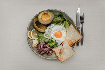 早餐包括煎鸡蛋，将其放在新鲜蔬菜、米浆果、面包和大蒜上，鳄梨切成两半，柠檬片放在陶瓷盘上。