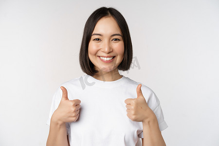 故障反馈摄影照片_快乐的韩国年轻女性竖起大拇指微笑，给出积极反馈，推荐 smth，站在白色背景上