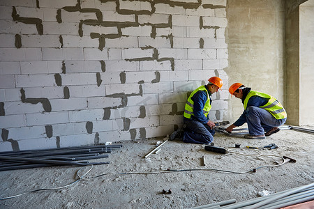 两名身着工作服、头戴安全帽的男性建筑工人正在使用现代工具在正在建设的建筑中安装塑料管