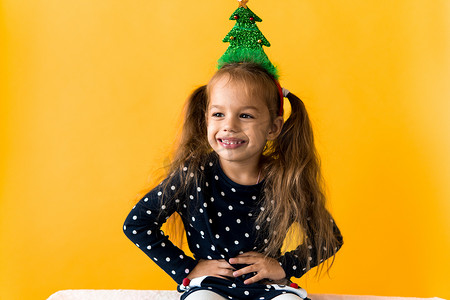 鹿黄色摄影照片_正面微笑的肖像令快乐的小女学生女孩圣诞树装饰在头部圆点连衣裙上，竖起大拇指橙色背景。