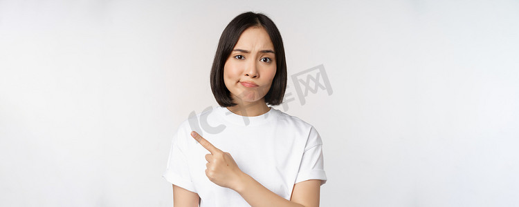 持怀疑态度的亚洲女孩穿着白色 T 恤，用失望的表情指着产品或标志，不喜欢并抱怨 smth，站在白色背景上