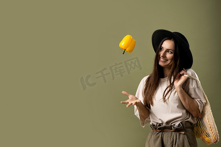 身穿米色 T 恤、头戴黑色帽子的生态友好型微笑女性单手向空中抛出一个黄色胡椒粉，肩上拿着装满杂货的可重复使用的棉质生态袋。