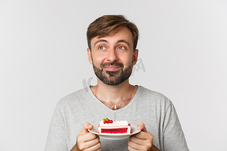 梦幻般的大胡子男人的特写镜头，微笑着庆祝生日，拿着点着蜡烛的蛋糕，许下生日愿望，站在白色背景上