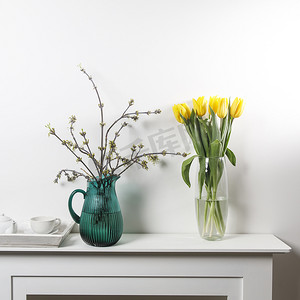 花束妈妈摄影照片_玻璃花瓶中的黄色郁金香花束和白桌上绿色玻璃罐中的美国枫树枝