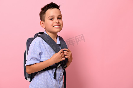 背书包的小学生摄影照片_一个微笑着露齿微笑的小学生的侧画像，他背上背着一个书包，背景是粉红色，有复制空间。