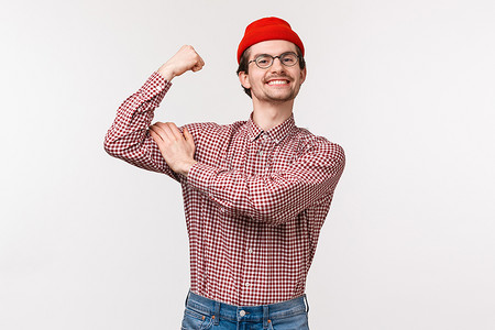 身穿红色帽子和眼镜的滑稽成年苗条男人的腰上画像表现得酷酷而强硬，弯曲的二头肌显示出他有多么强壮，骄傲而满意的微笑，在健身房锻炼，站立的白色背景