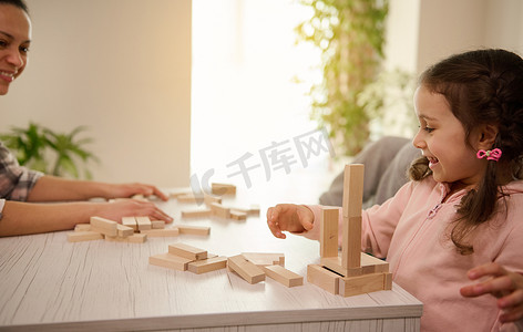 漂亮的孩子，穿着粉色运动衫的可爱小女孩和妈妈玩棋盘游戏，用木砖和木块搭建木结构，露出露齿的笑容，享受她的教育消遣